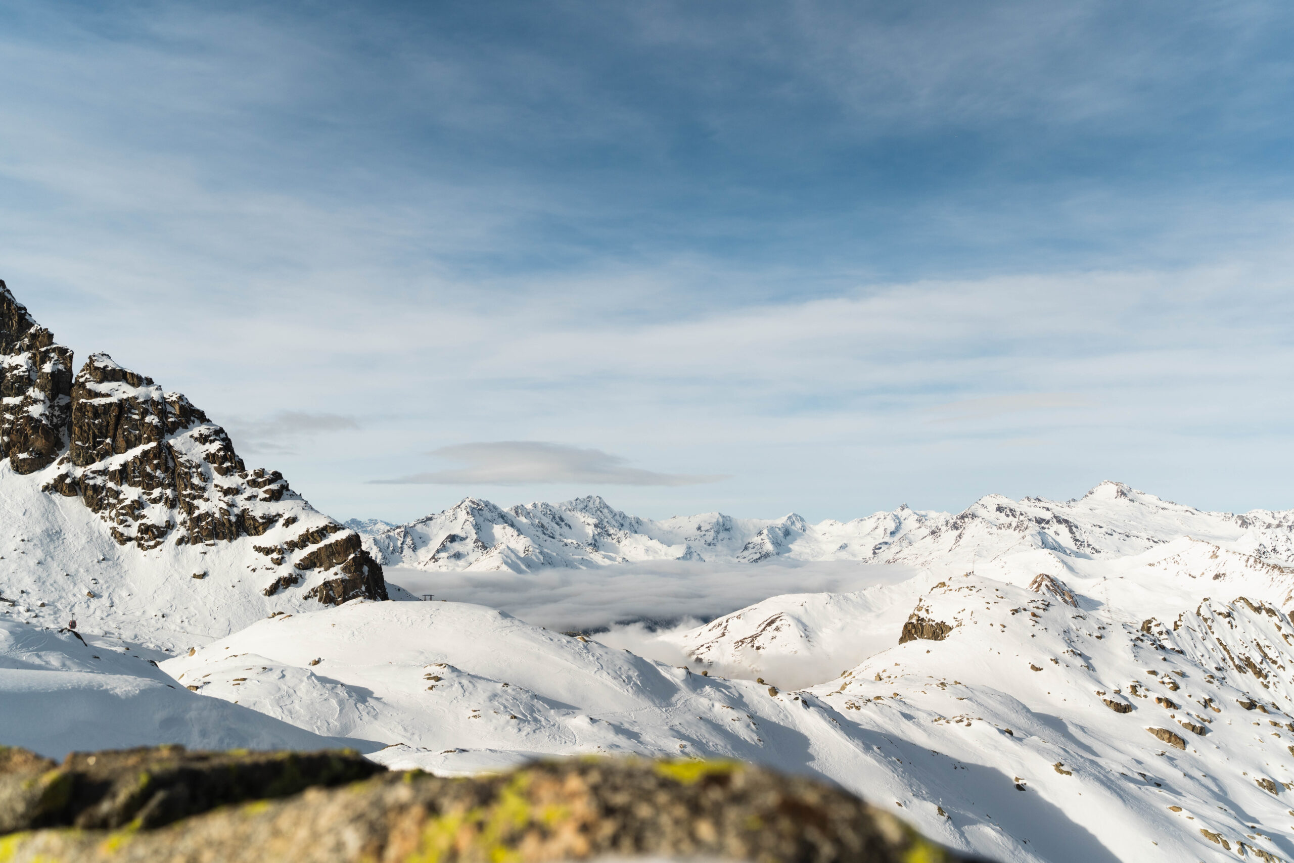 Snow-covered peaks of Presena Glacier with clouds below the mountains under a clear blue sky. The sunlight highlights the rugged terrain and winter landscape