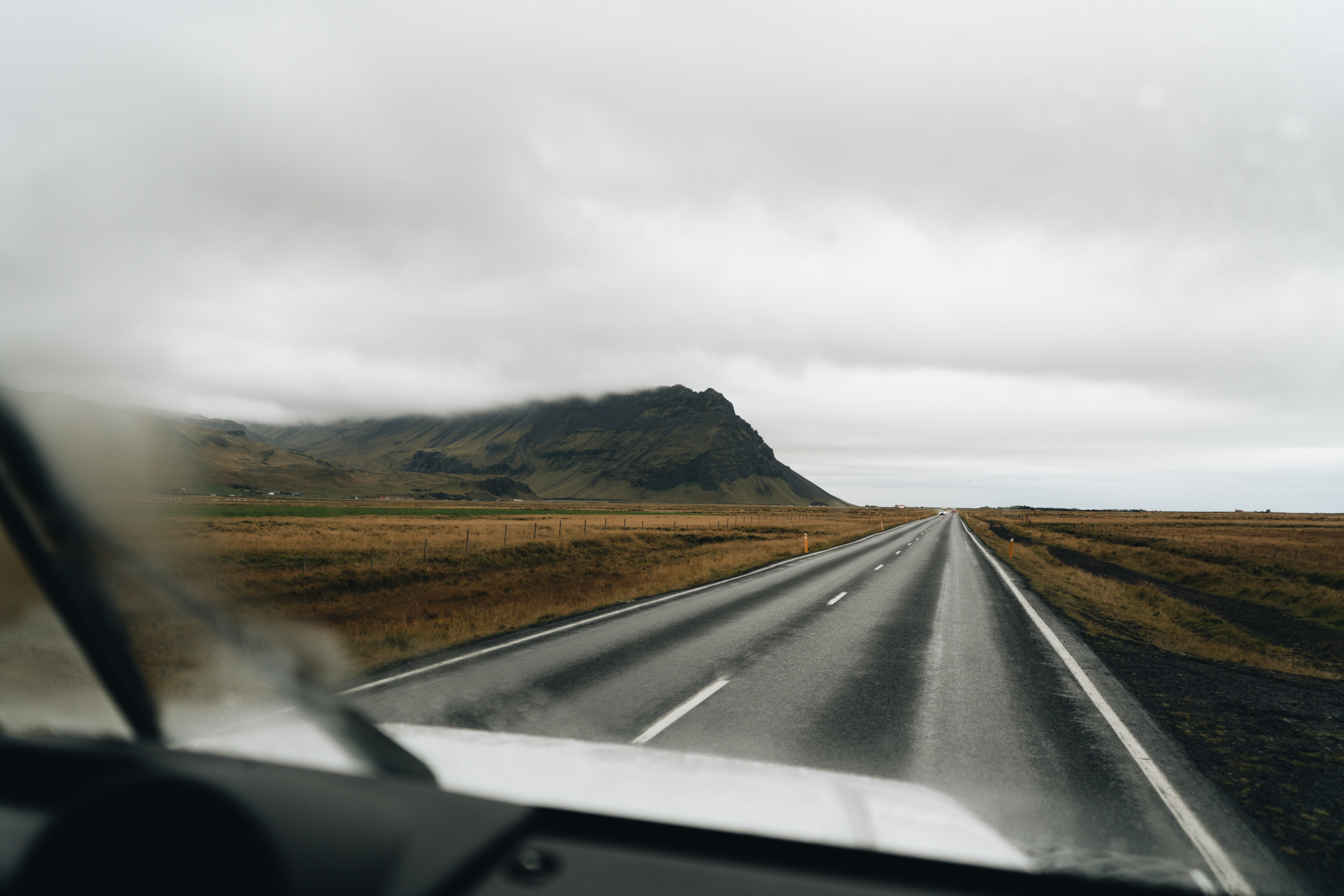 View from a car windshield of a misty, overcast road in Thórsmörk, Southern Iceland, surrounded by vast grassy fields and rugged hills under gray skies, 2023