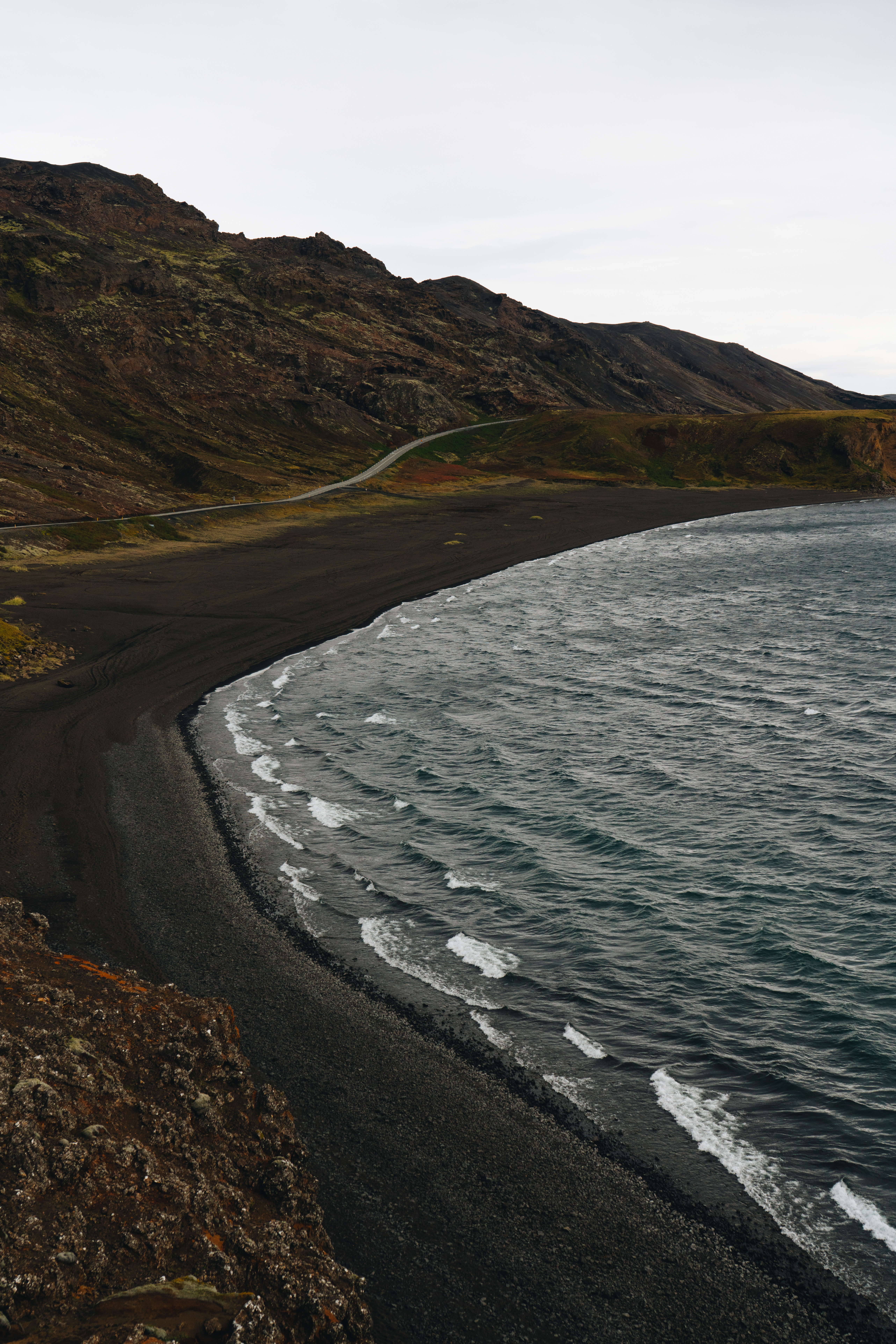 Winding coastal road along black sand beach in Iceland, showcasing dramatic contrast between dark volcanic sand and rolling waves against rugged mountain landscape