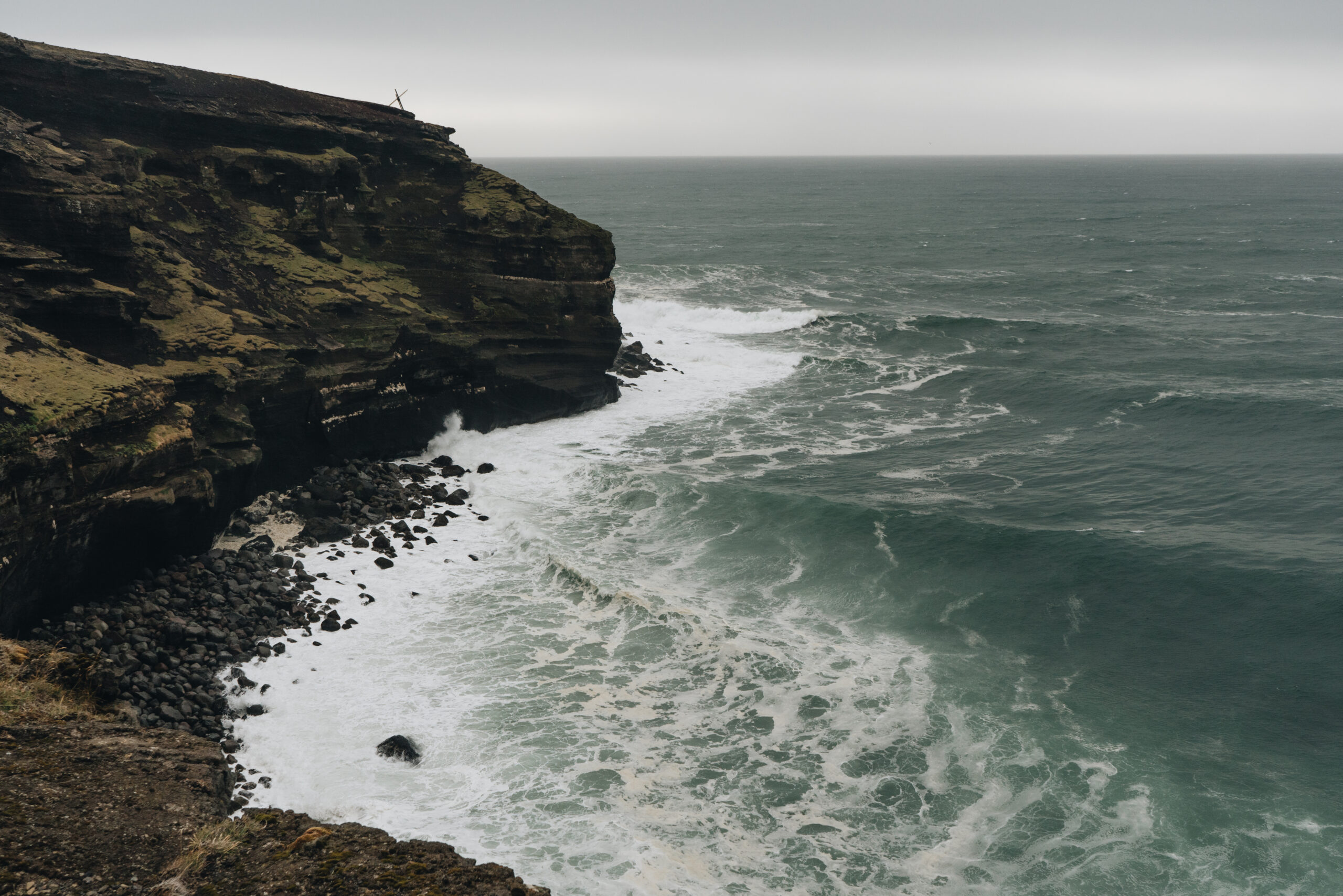 Dramatic coastal cliffs at Krísuvik Iceland with turbulent emerald-green ocean waves crashing against dark volcanic rocks, featuring layered cliff formations and white sea spray against a moody sky