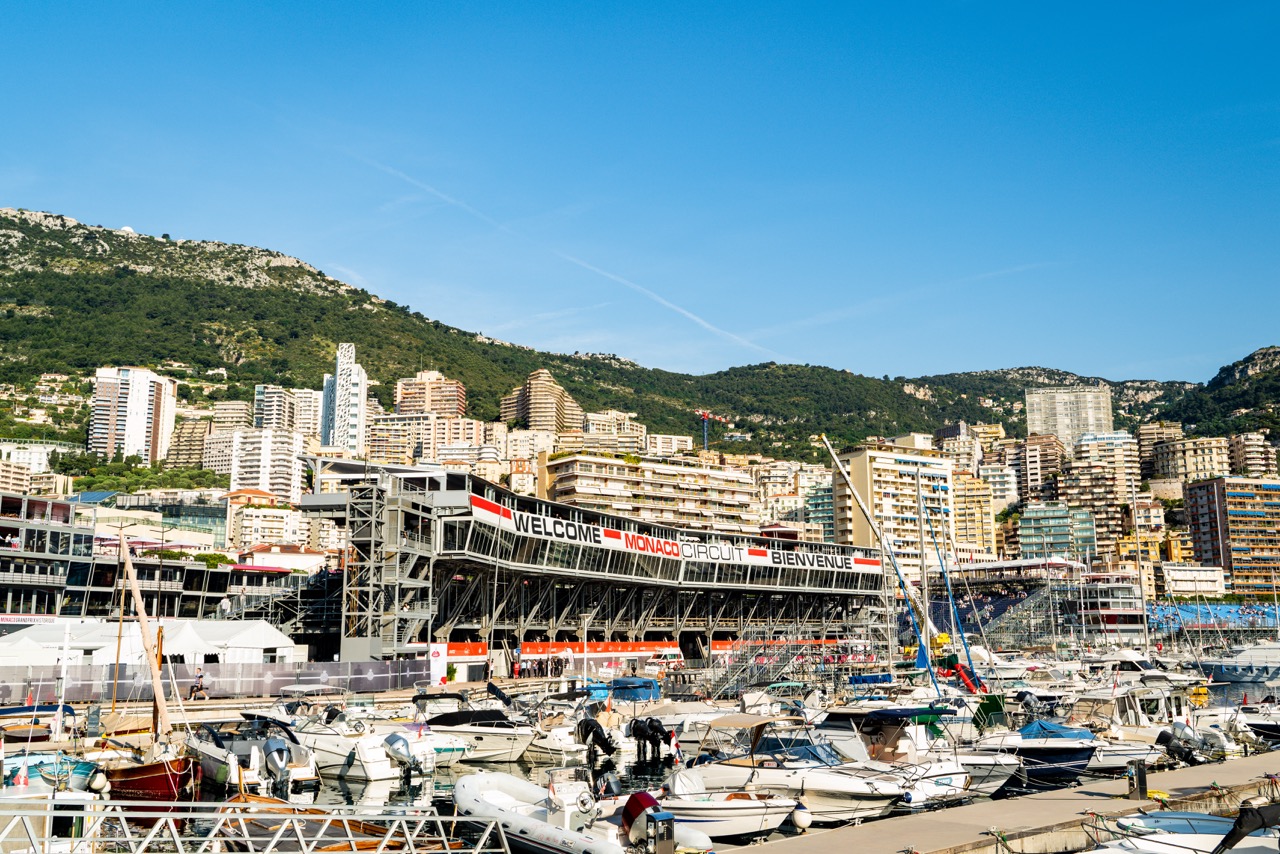 Wide view of the Monaco Circuit’s harbor area, showcasing luxury yachts, grandstands, and the ‘Welcome Monaco Circuit Bienvenue’ sign. The backdrop features densely packed buildings and lush hills under a clear blue sky.