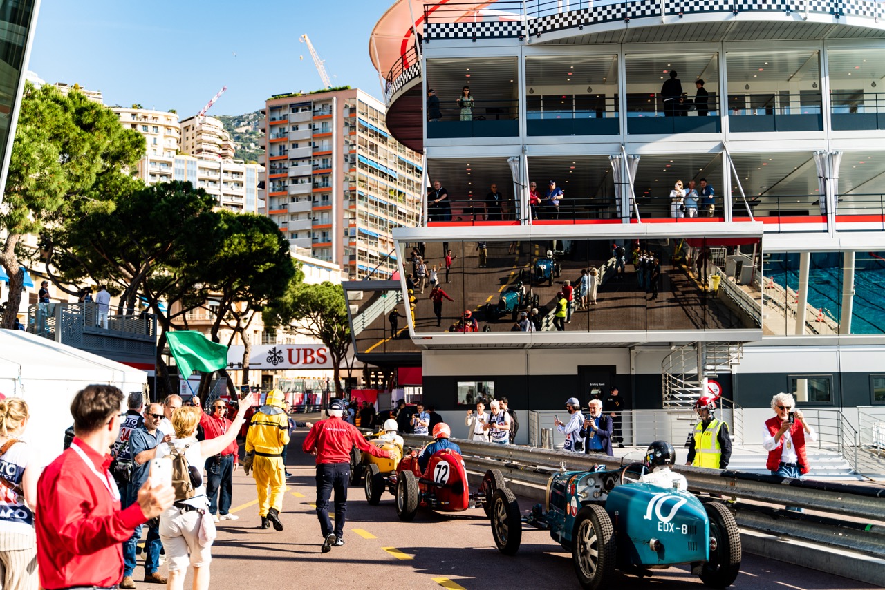 Vintage race cars lined up in a Monaco pit lane, surrounded by spectators, marshals, and event staff. The setting includes a large, modern building with balconies, reflecting the vibrant race activity