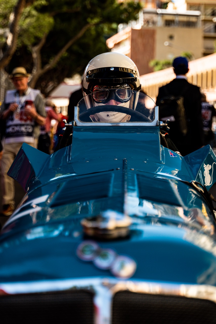 Close-up of a driver in a vintage blue race car, wearing a cream helmet and goggles. The focus is on the driver’s intense expression behind the wheel, with a blurred crowd and trees in the background
