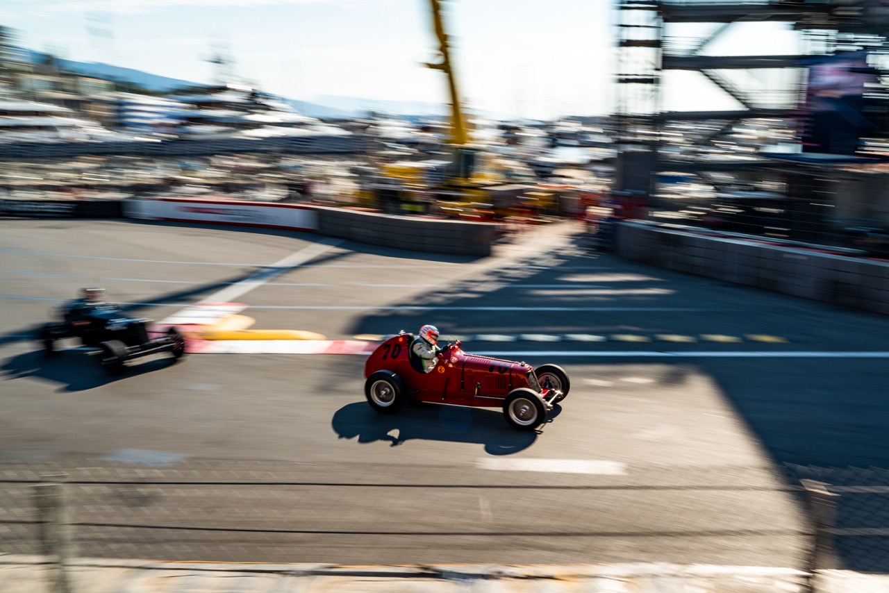 Two vintage race cars speeding through a sharp corner on a racetrack. The red car with the number 70 leads, while a black car follows. Motion blur highlights the cars’ speed, with cranes, barriers, and yachts visible in the background
