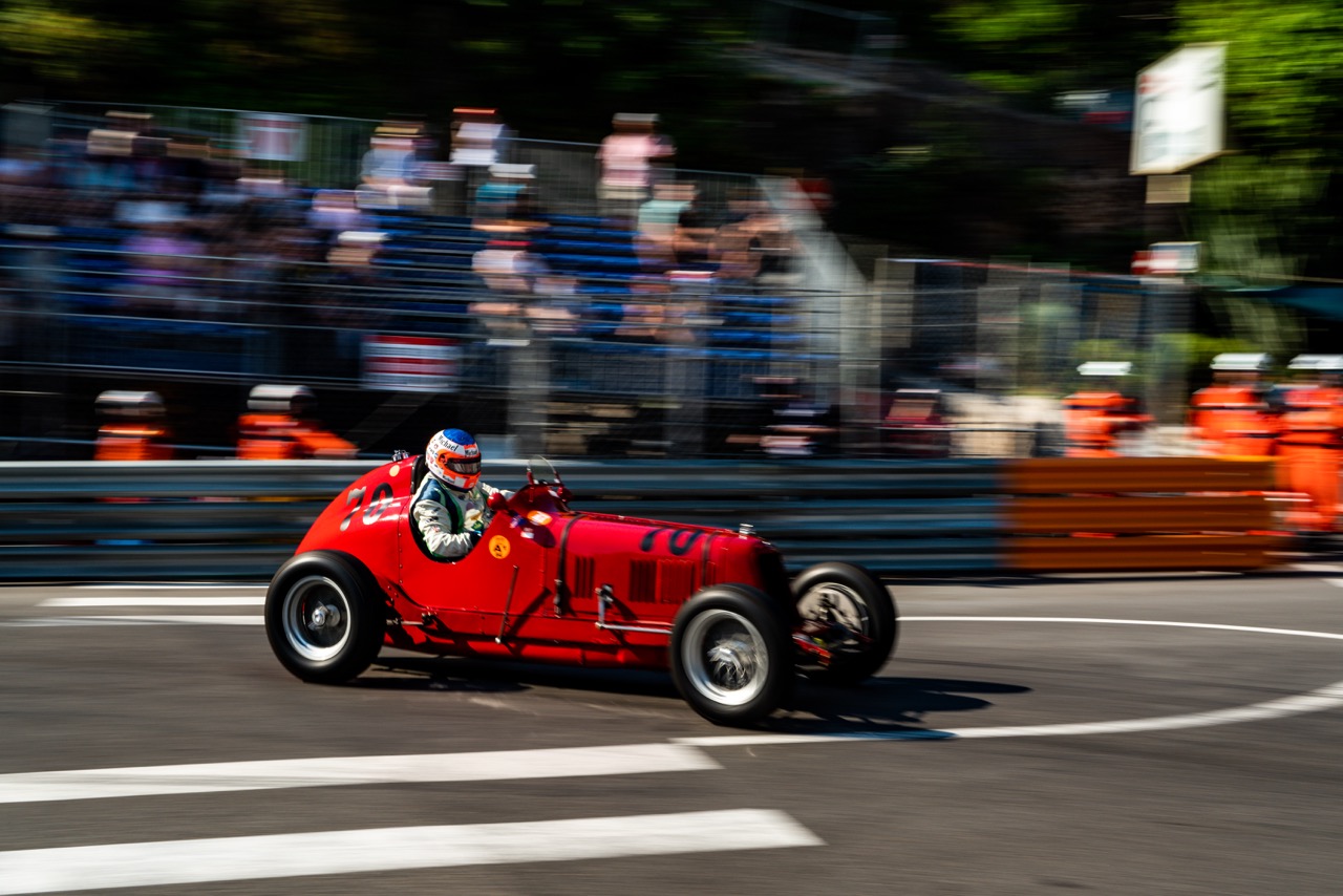 Close-up of a red vintage race car with the number 70, driven by a racer wearing a blue helmet. The car is navigating a curve on the racetrack with motion blur in the background, showcasing the crowd and safety barriers.