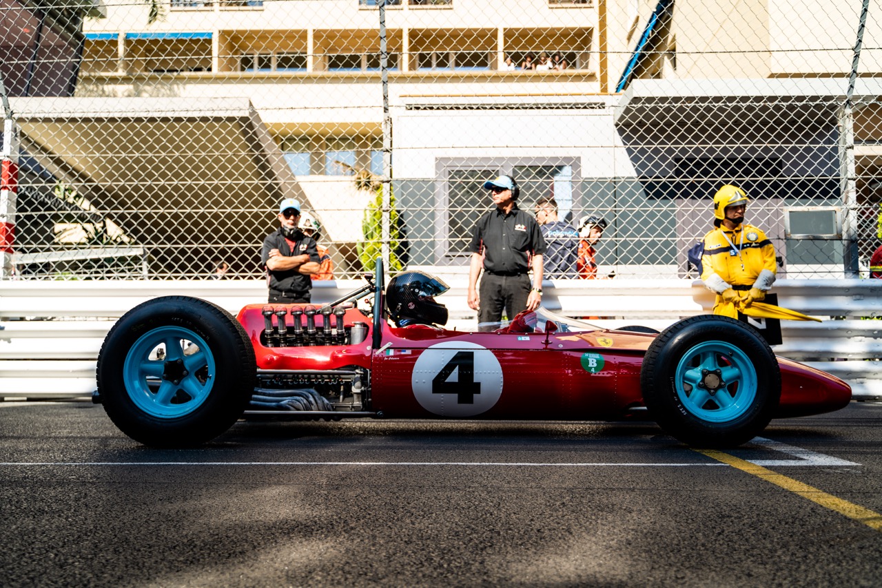 Side view of a vintage red Formula 1 car with the number 4 on its body, parked on the track. Crew members and a safety marshal in a yellow uniform stand behind the car near a chain-link fence, with modern buildings in the background