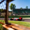 _CLC4786 A vintage Formula 1 car with Marlboro sponsorship, captured at high speed on a racetrack. The car is framed artistically through a wire fence, emphasizing the dynamic motion of the vehicle with blurred surroundings. The background includes green spectator stands under a clear blue sky