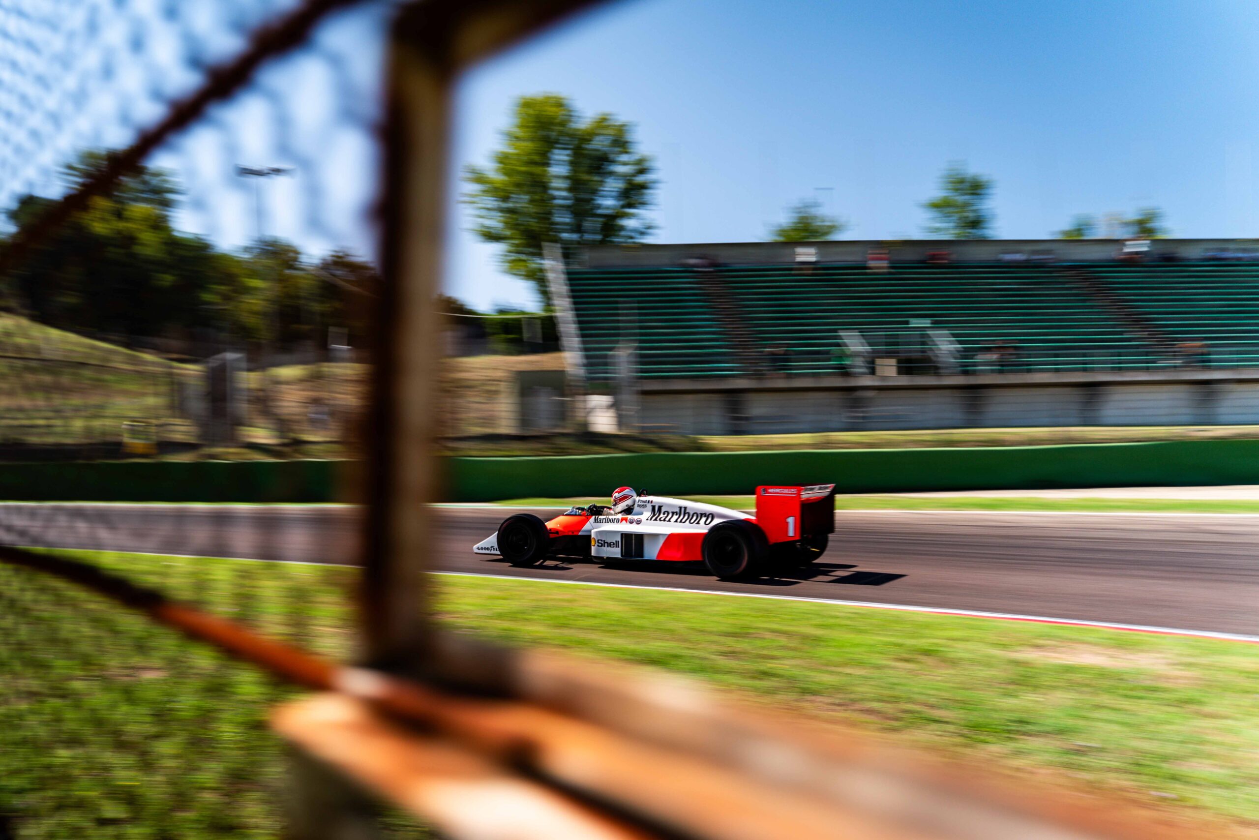 A vintage Formula 1 car with Marlboro sponsorship, captured at high speed on a racetrack. The car is framed artistically through a wire fence, emphasizing the dynamic motion of the vehicle with blurred surroundings. The background includes green spectator stands under a clear blue sky