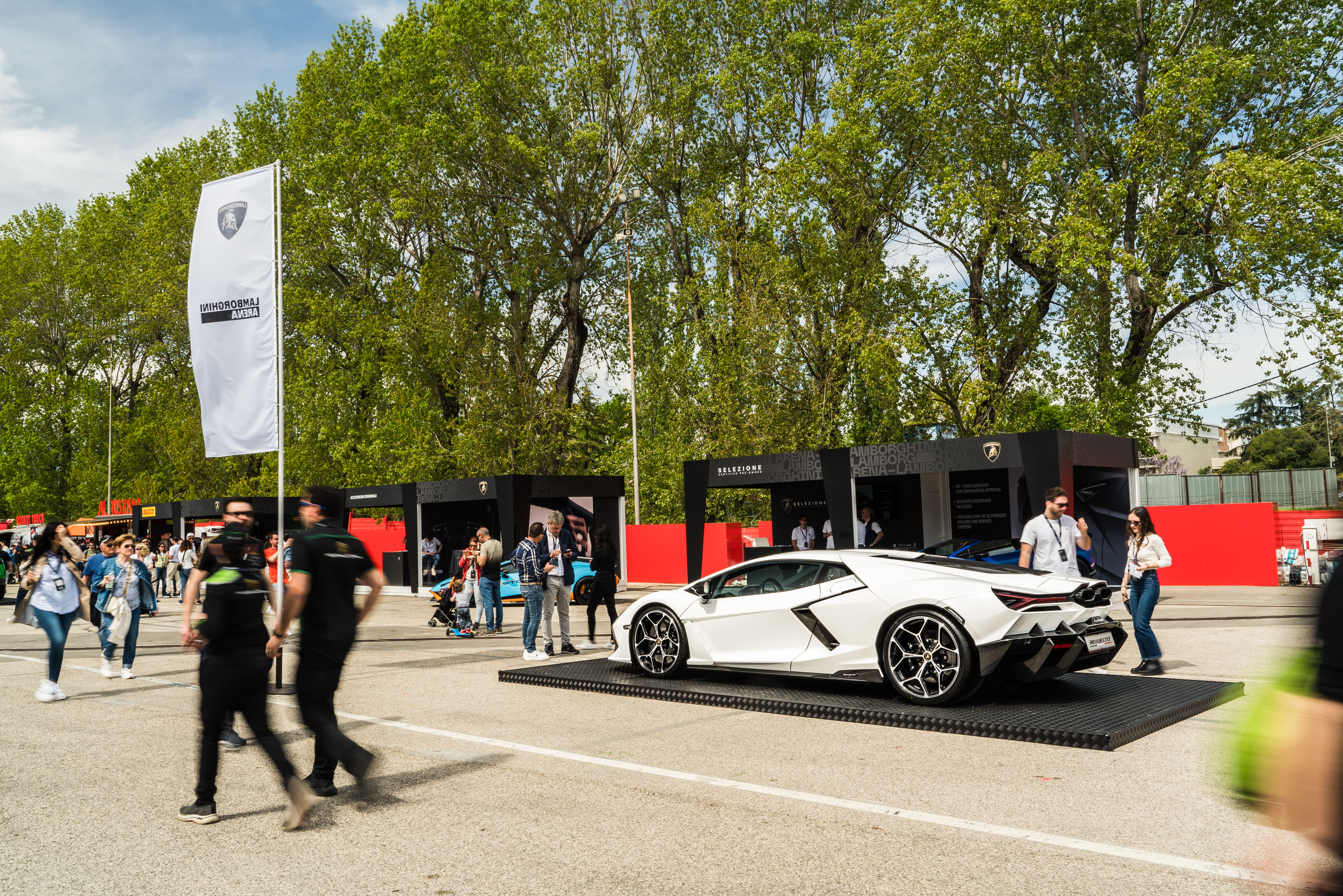 A white Lamborghini on display at a public event area, surrounded by people and branding