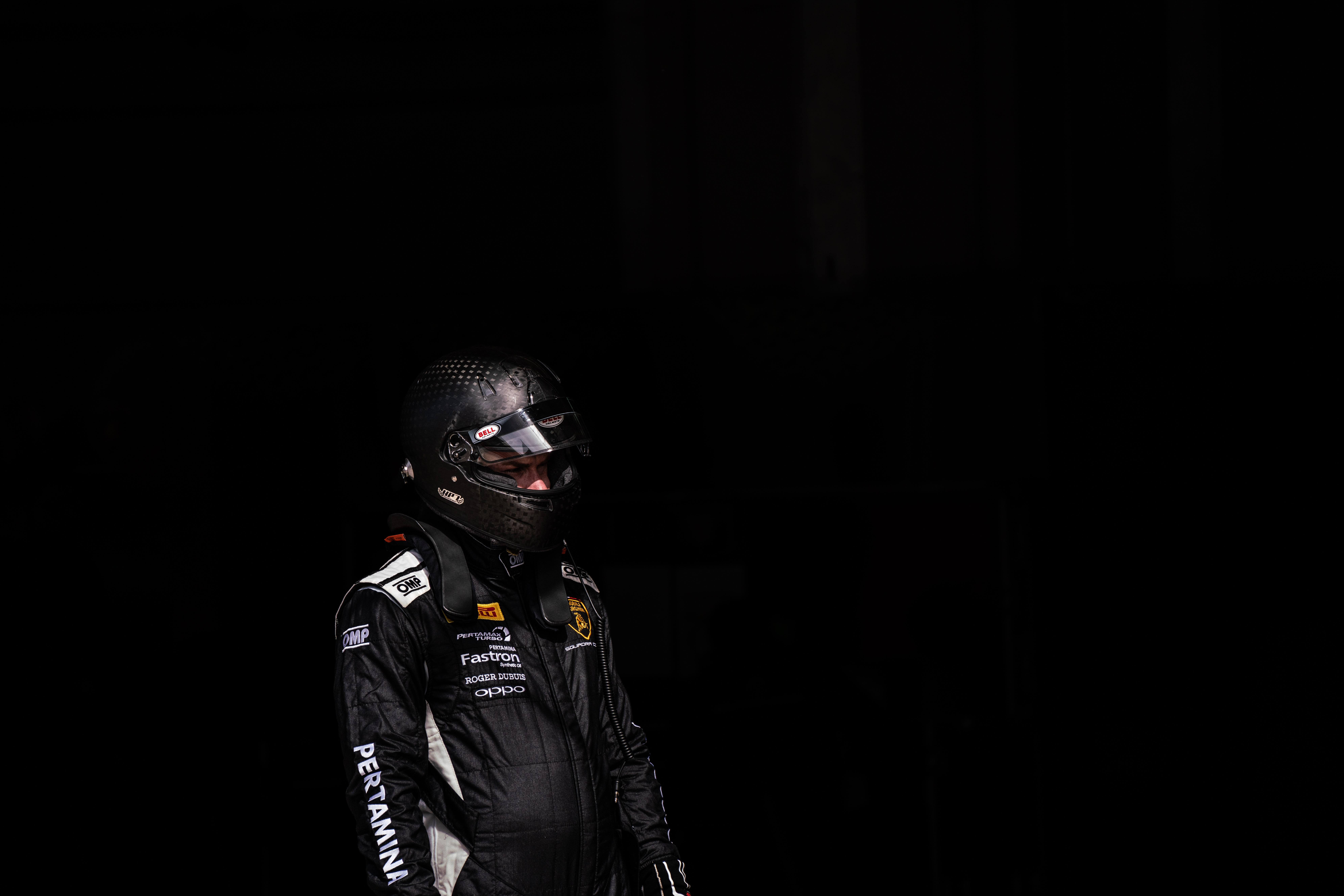 Driver in black racing gear with a helmet, standing in a dark pit area, ready for the race