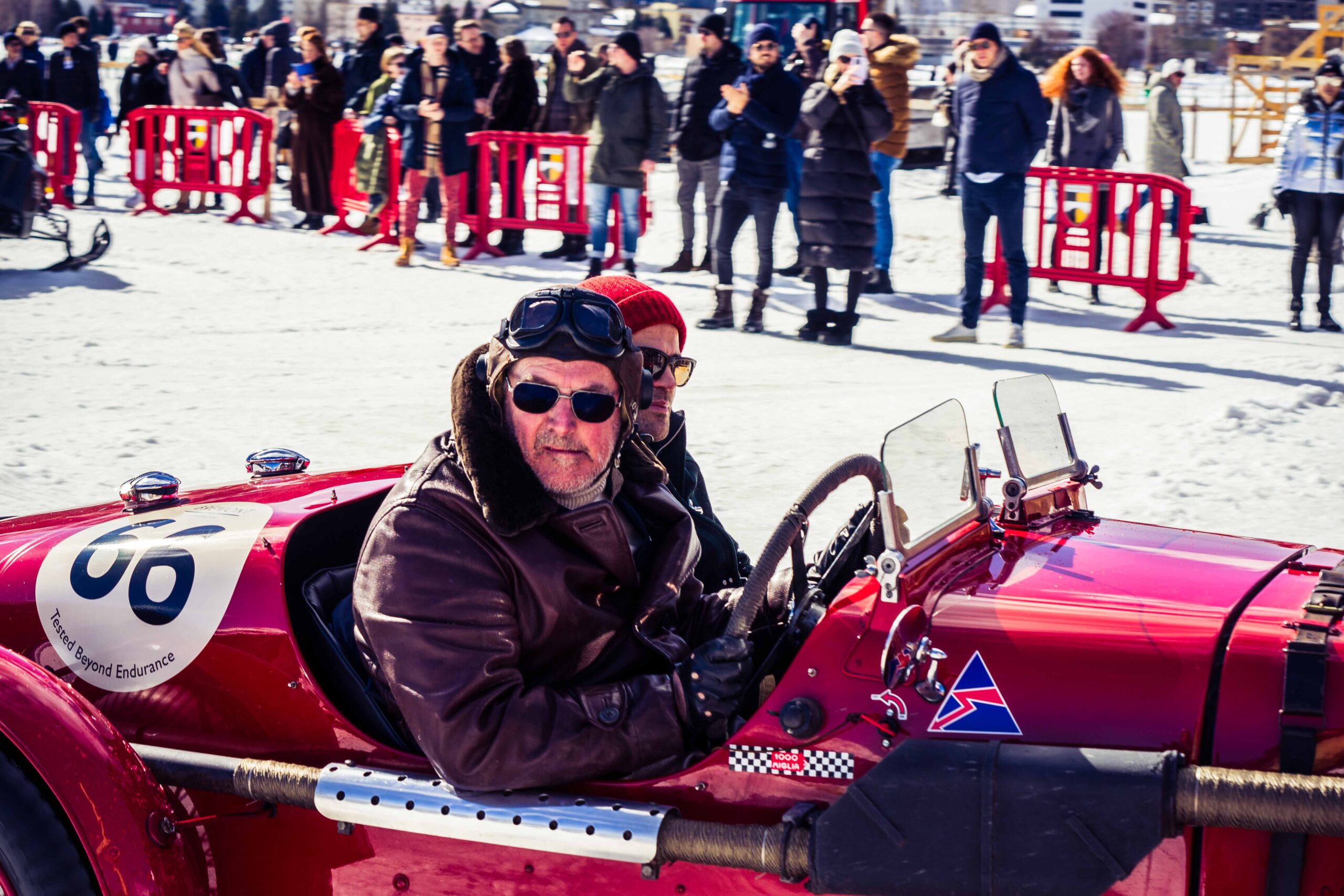 Driver wearing vintage goggles and leather jacket in a red classic race car, surrounded by spectators at The I.C.E. St. Moritz 2023
