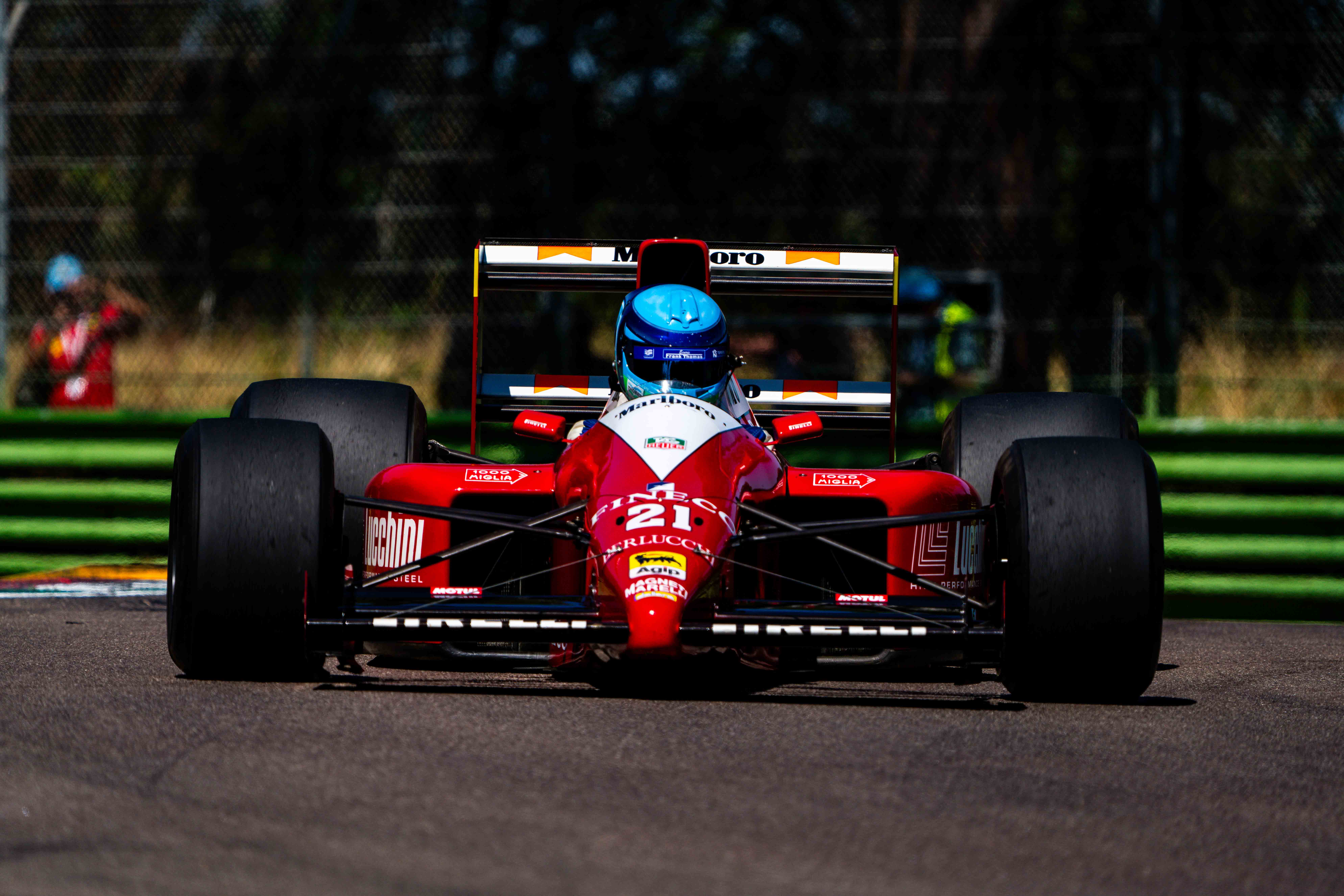 A front-facing shot of a vintage red Formula 1 car with a vibrant blue-helmeted driver, captured sharply as it speeds down a racetrack. The car features classic sponsorship decals, with a blurred green and brown background adding depth to the action scene