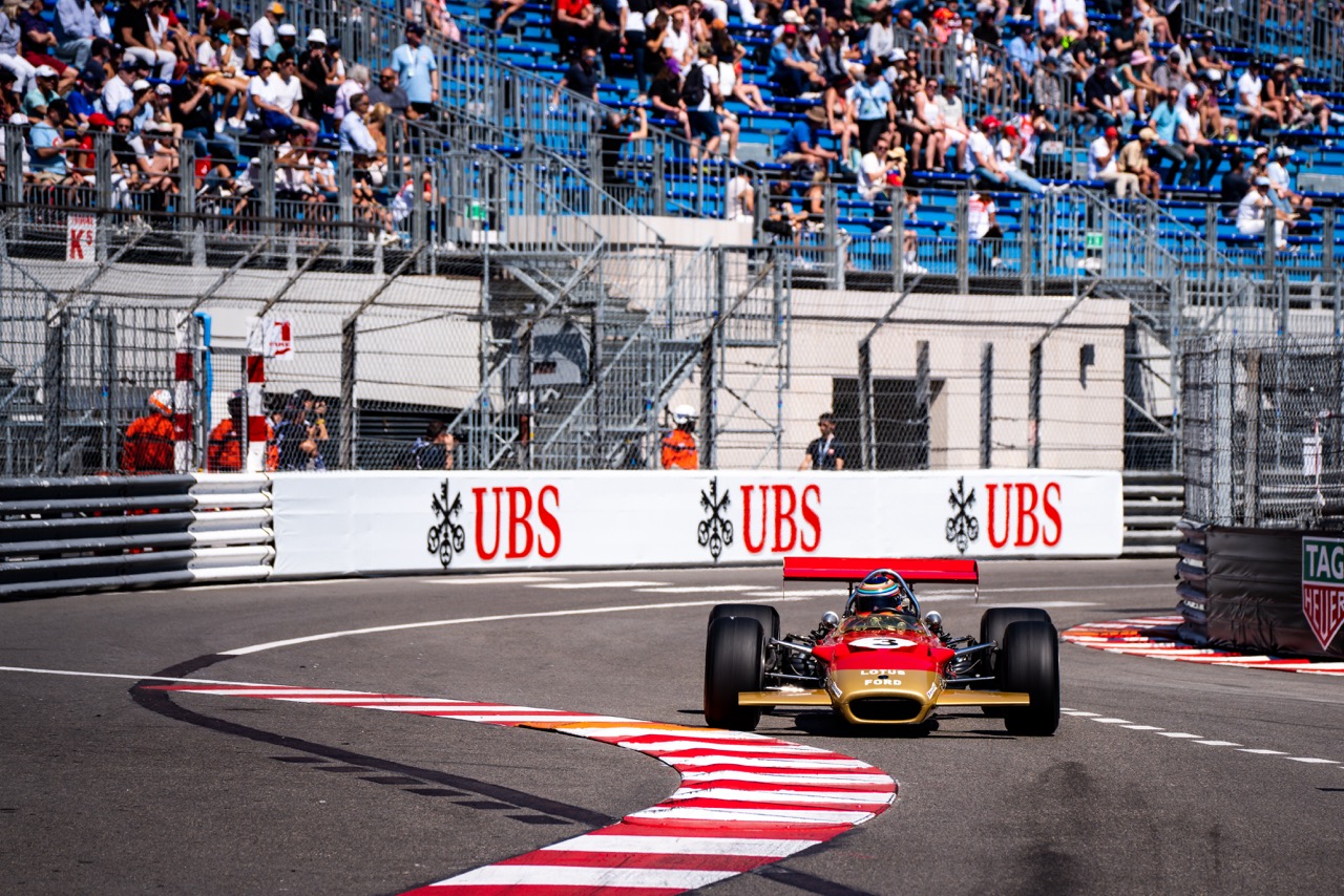 A gold and red vintage Lotus Ford Formula 1 car taking a corner on a racetrack. The driver is visible in the cockpit, wearing a helmet, while a grandstand filled with spectators