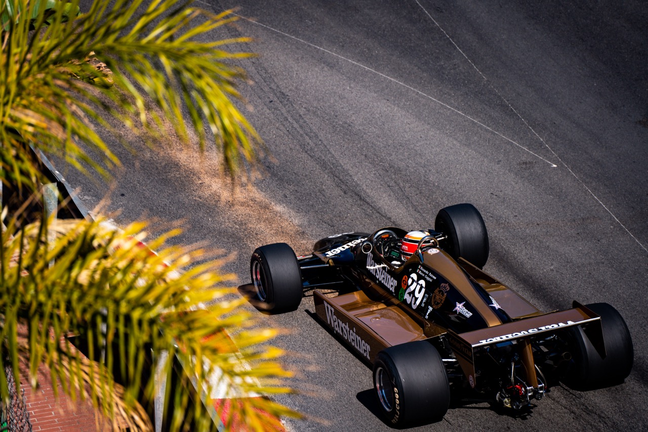Top view of a vintage Formula 1 car racing through a corner on a winding asphalt track. Palm leaves frame the foreground, highlighting a sunny, outdoor race setting