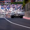 Rear view of a classic Formula 1 car on a street circuit, with a waving driver celebrating amidst barriers and grandstands full of fans. Luxury yachts and a vibrant crowd in the background capture the atmosphere of the Monaco Historique Grand Prix