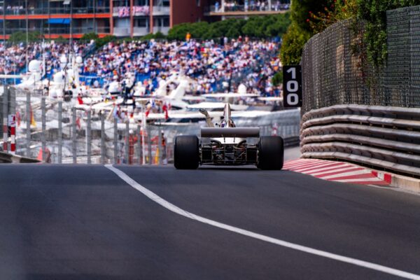 Rear view of a classic Formula 1 car on a street circuit, with a waving driver celebrating amidst barriers and grandstands full of fans. Luxury yachts and a vibrant crowd in the background capture the atmosphere of the Monaco Historique Grand Prix