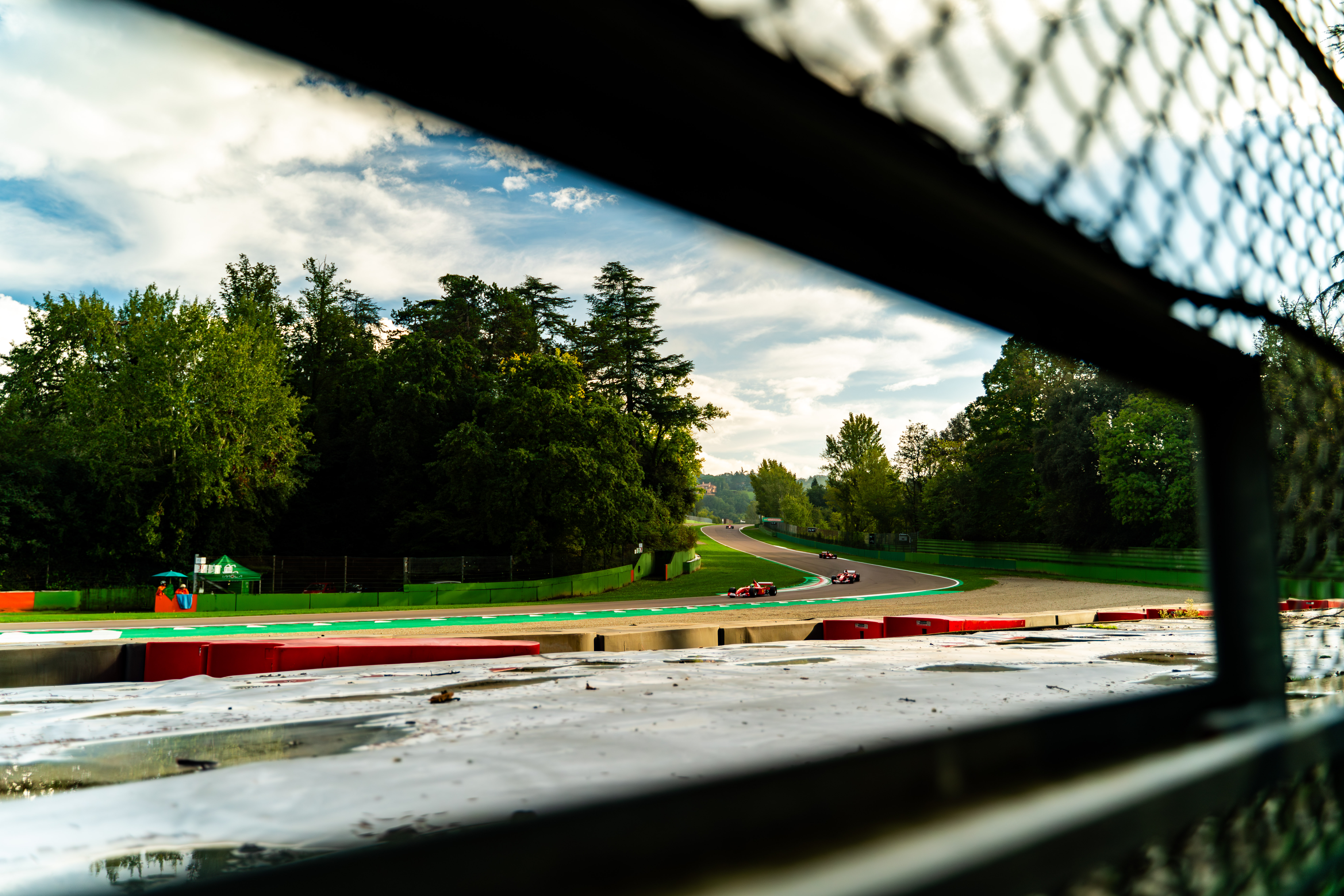 View of the Imola racetrack through a fence, showcasing a scenic section with Ferrari cars on a curving road surrounded by trees