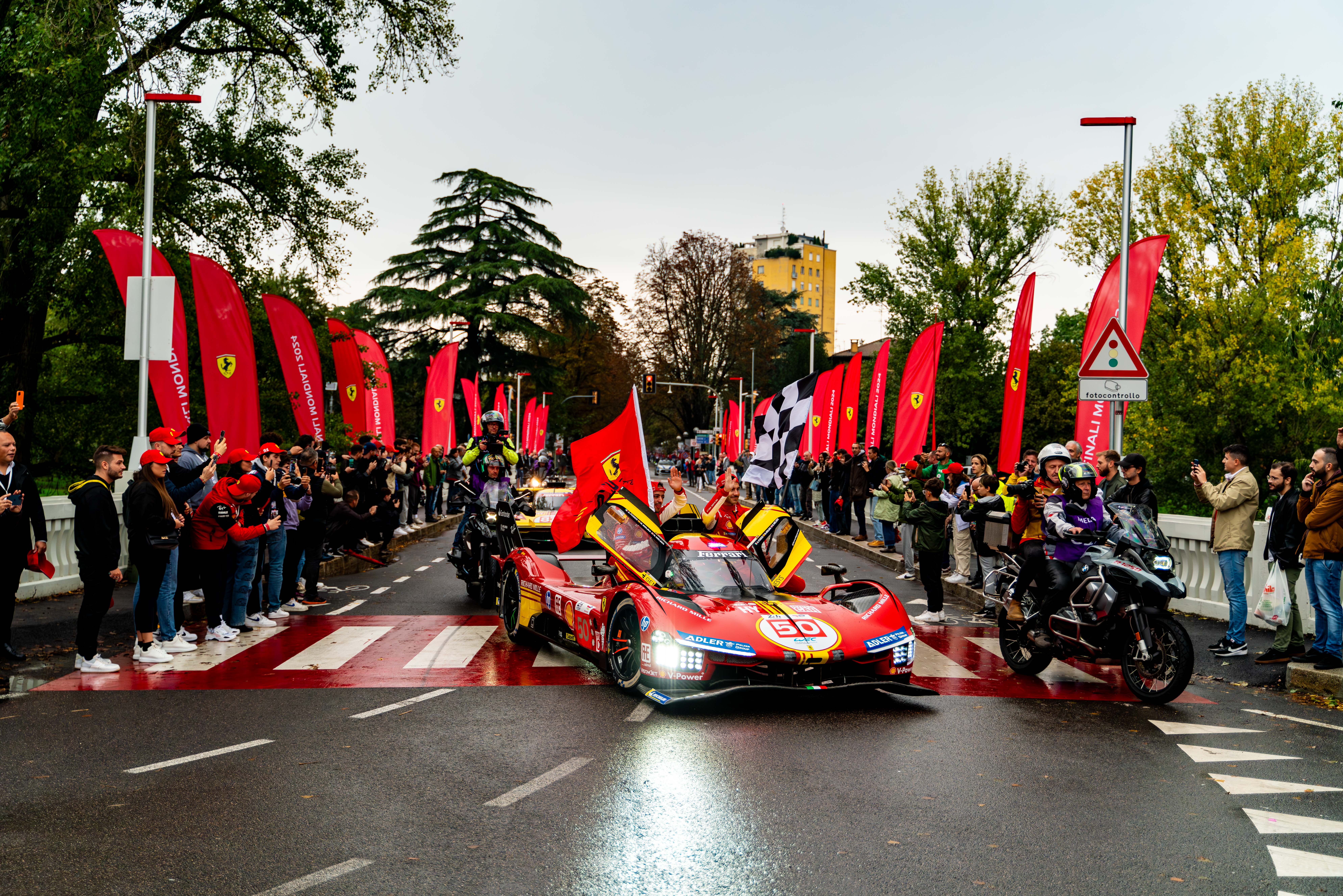 Ferrari car leading a parade on a city street, surrounded by fans waving red flags