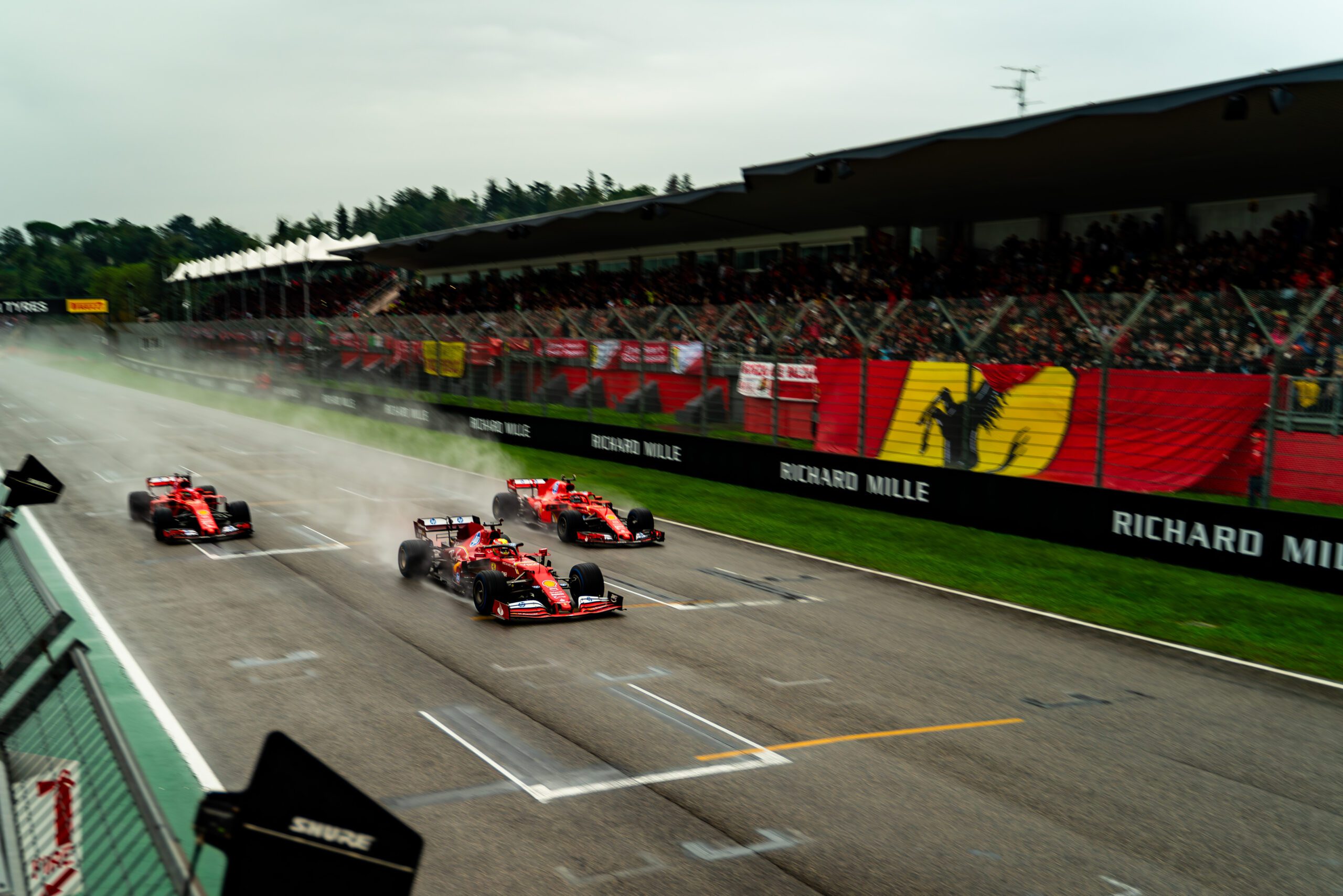 Ferrari Formula 1 cars accelerating side by side on a rain-soaked race track with grandstands in the background