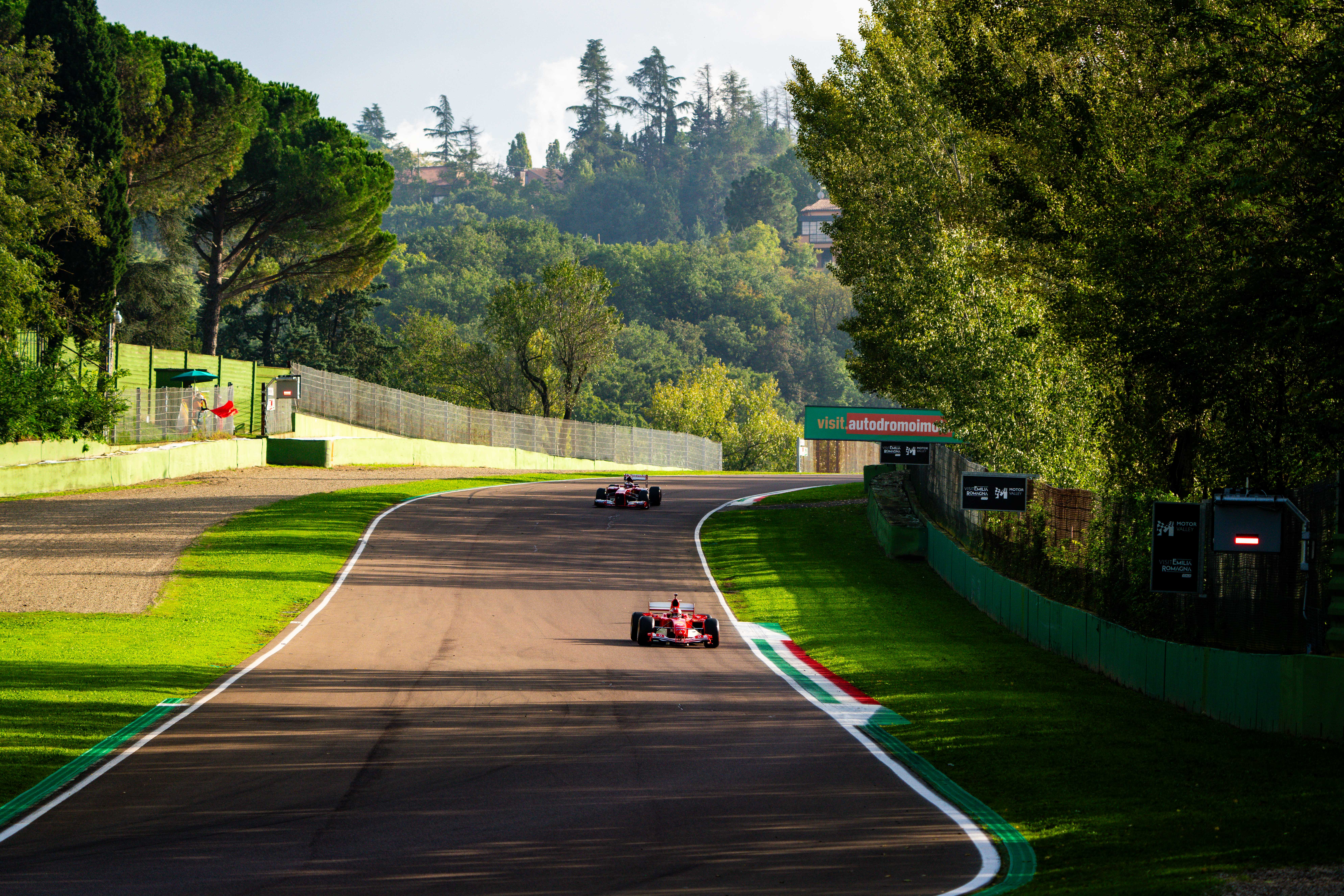 Two Ferrari Formula 1 cars driving through a forested section of the Imola racetrack, framed by lush greenery and a sunny sky