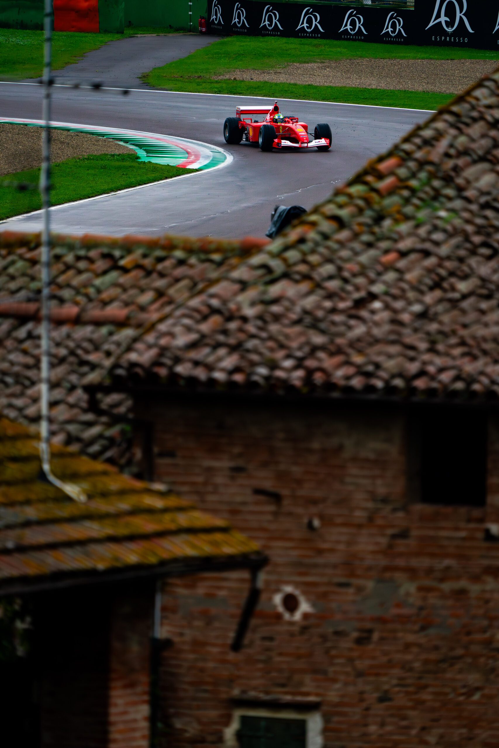 Distant view of a Ferrari Formula 1 car passing through a corner of the Imola circuit, seen over the rooftops of historic buildings